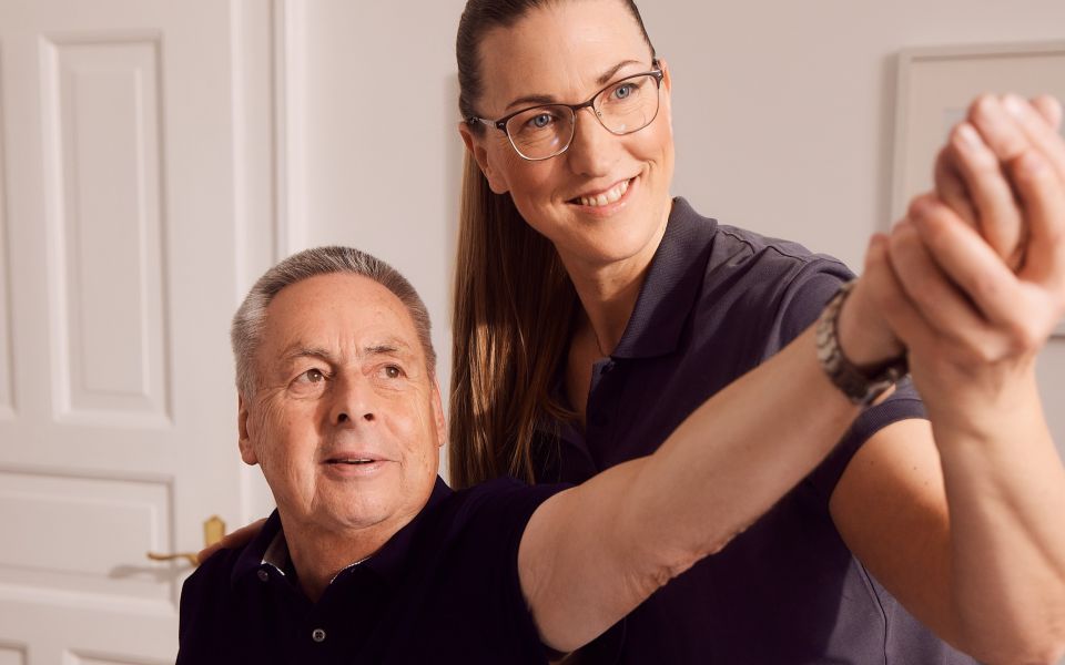 Physiotherapeutin hilft älterem Mann bei Armübungen im Raum.